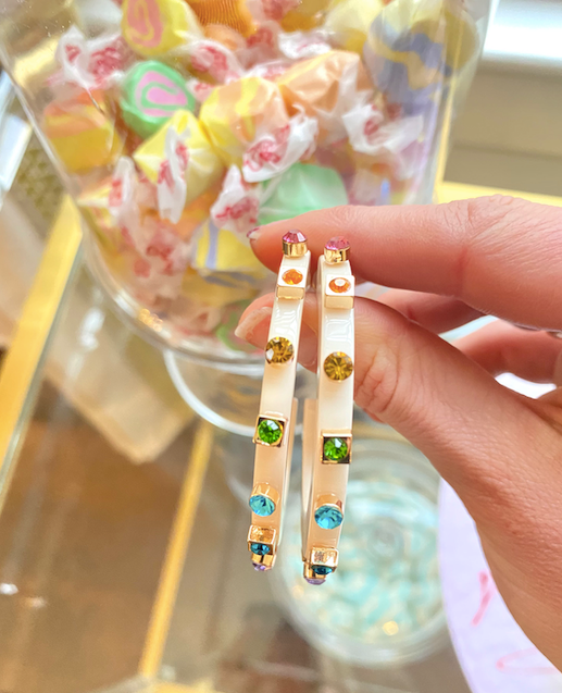 Hand holding a pair of cream-colored hoop earrings, embellished with colorful gemstones, against a background of pastel-wrapped candies in a glass jar.