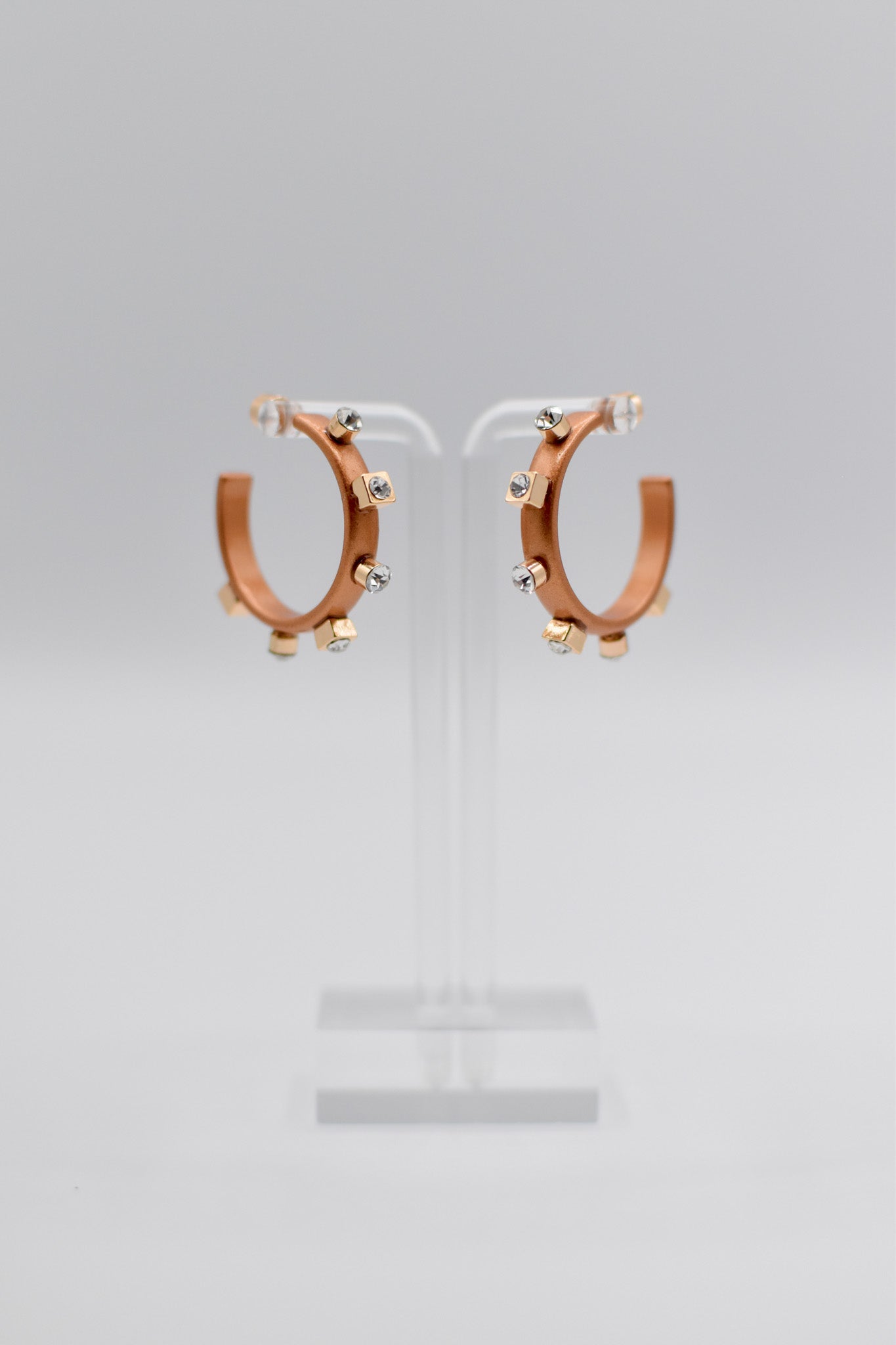 Two copper metal hoop earrings are displayed on a transparent stand, adorned with small crystals and gold metallic studs along their outer edge.