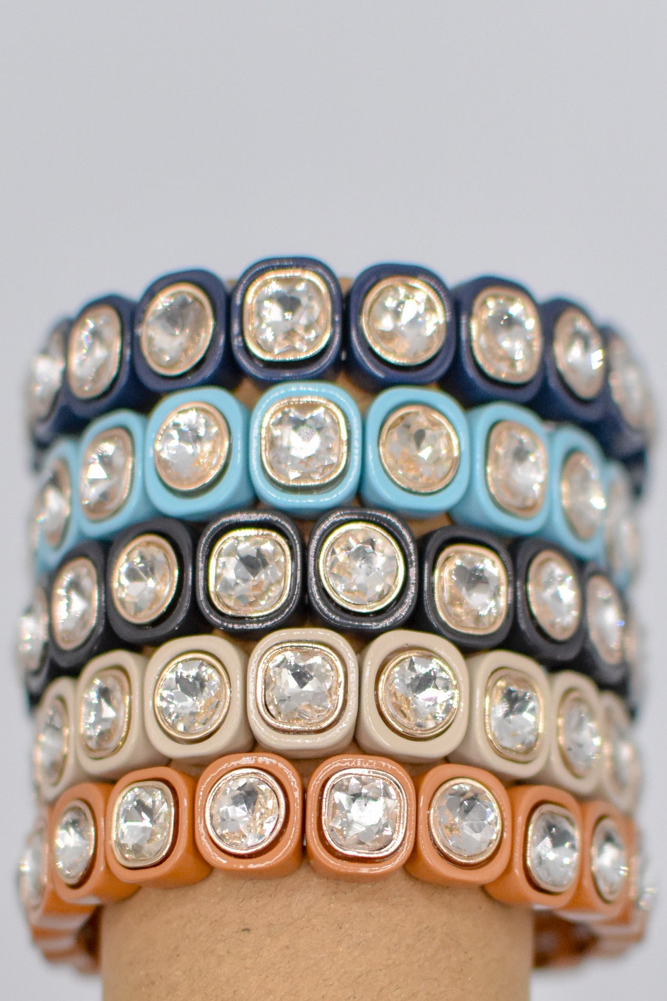A stack of colorful bracelets features multiple rows of square-shaped settings, each holding a sparkling crystal, arranged neatly in a vibrant display.