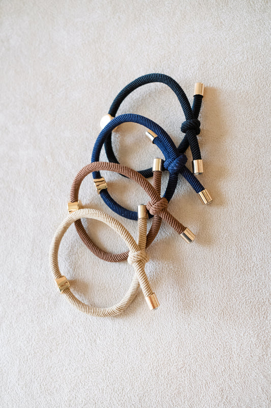 Set of four thick elastic hair ties in neutral and dark tones, including beige, brown, navy, and black, each with gold accents and knotted designs.