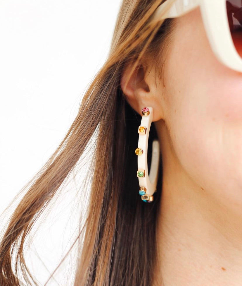 A close-up shot features a cream-colored hoop earring adorned with small, colorful gems along the outer edge, worn by someone with flowing brown hair.