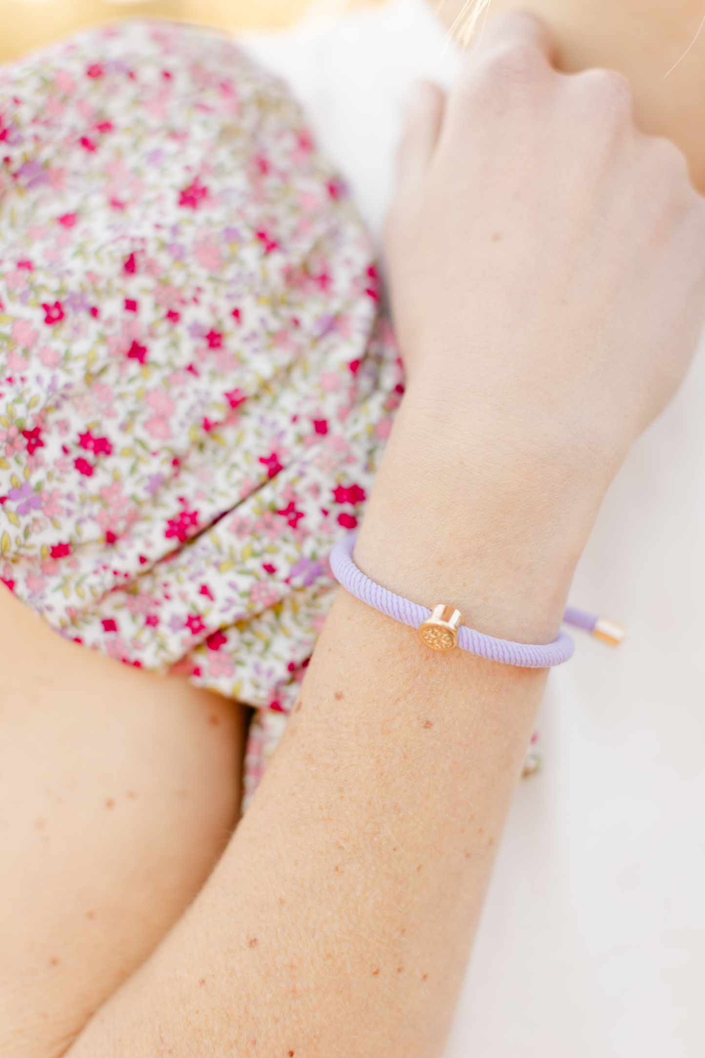 A close-up shot of a lavender elastic hair tie with gold accents, styled as a bracelet on the wrist, paired with a floral-patterned top.
