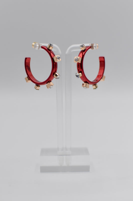 A pair of glossy, petite red metallic metal hoops, each adorned with six small crystal jewels on gold studs, are mounted on a transparent stand.