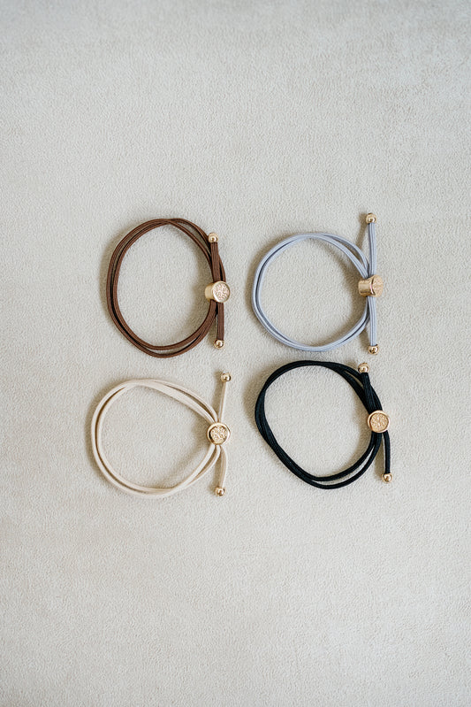 Four neutral-colored elastic hair ties in beige, brown, grey, and black, arranged in a grid with gold accents, showcasing a sleek, minimalist design.