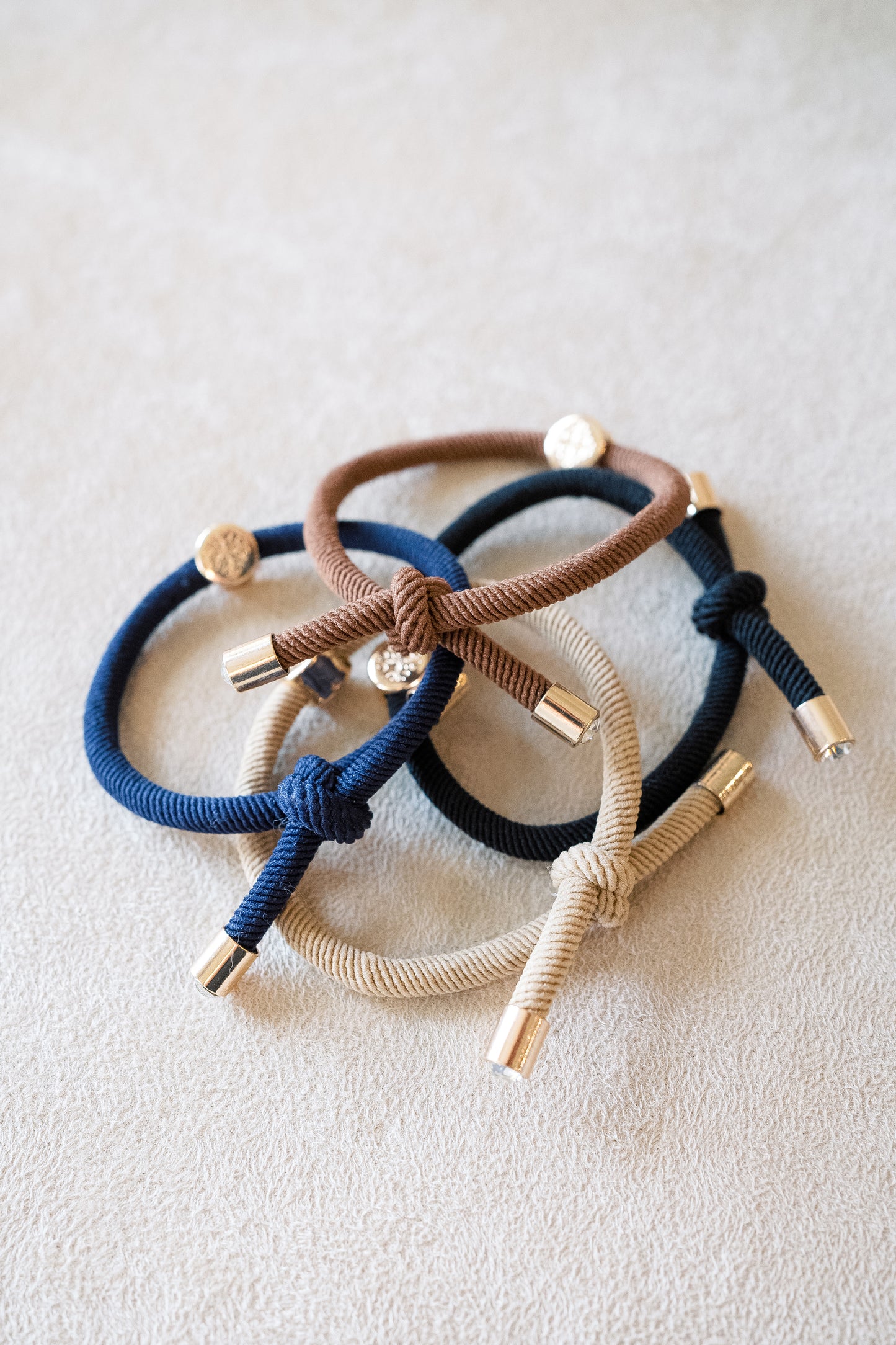 Set of four thick and textured elastic hair ties in neutral tones like beige, brown, navy, and black, each featuring gold accents and knotted designs.