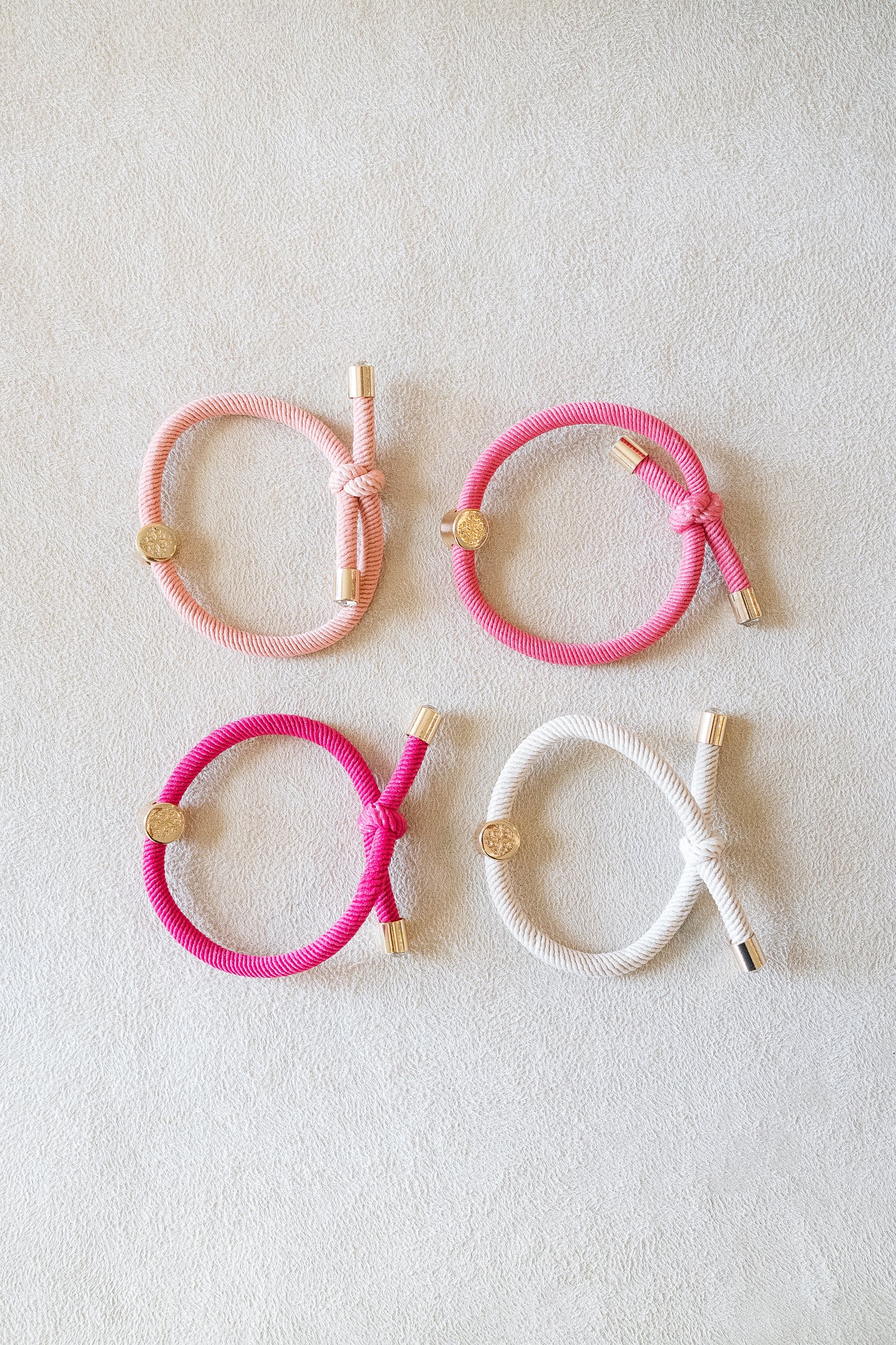 Four elastic hair ties in soft pink, vibrant pink, and white, arranged neatly in a grid on a light beige surface, each accented with shiny gold details.