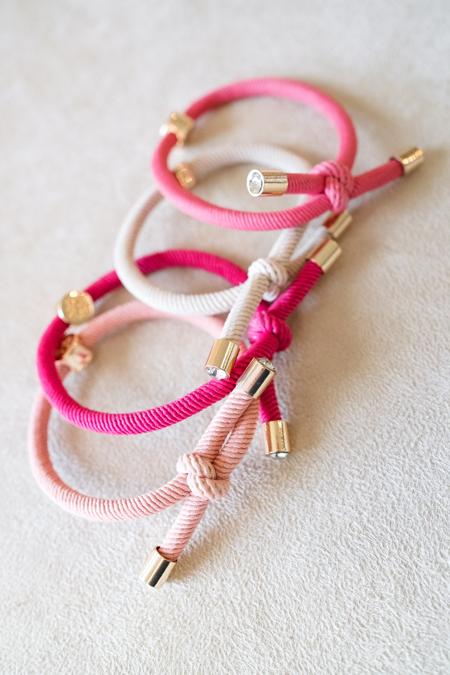 Four hair ties in shades of pink and beige with gold accents, arranged in a stack on a beige surface, showcasing their vibrant colors and metallic details.