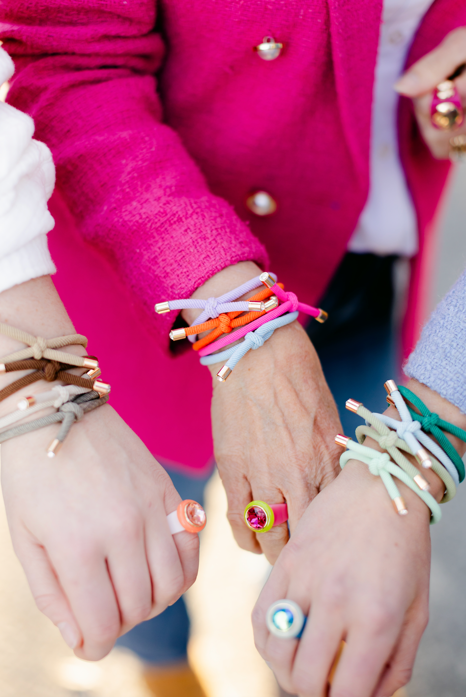Three models holding their wrists together wearing multi-colored jewel rings and stacks of four knotted hair ties in neutral, vibrant, and green color ways.