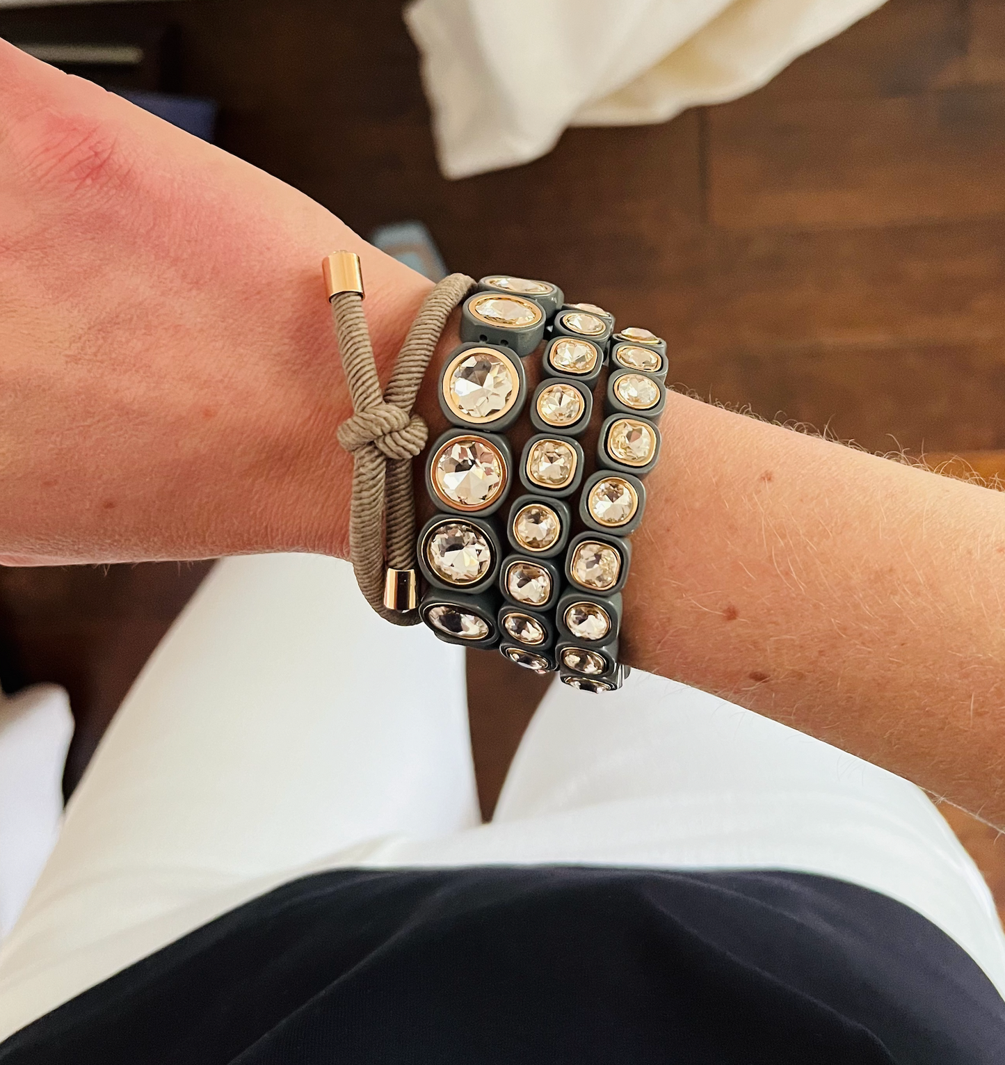 A wrist wearing a tan hair tie with gold accents and three different size sparkling crystal bands in light gray, featuring gold details around the stone.