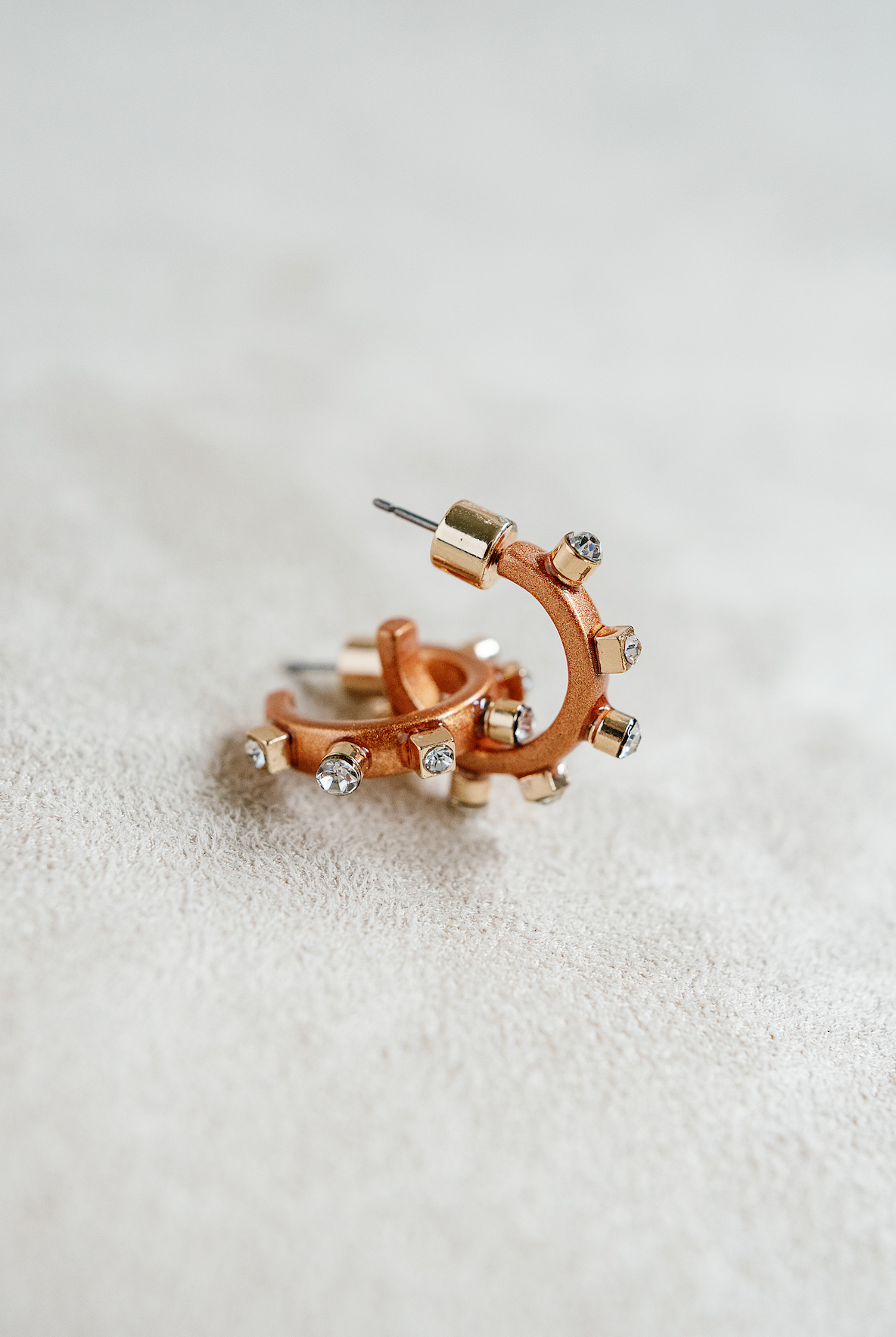 A pair of matte metal copper hoop earrings with gold accents, featuring studs around the rim and clear crystals on the post,