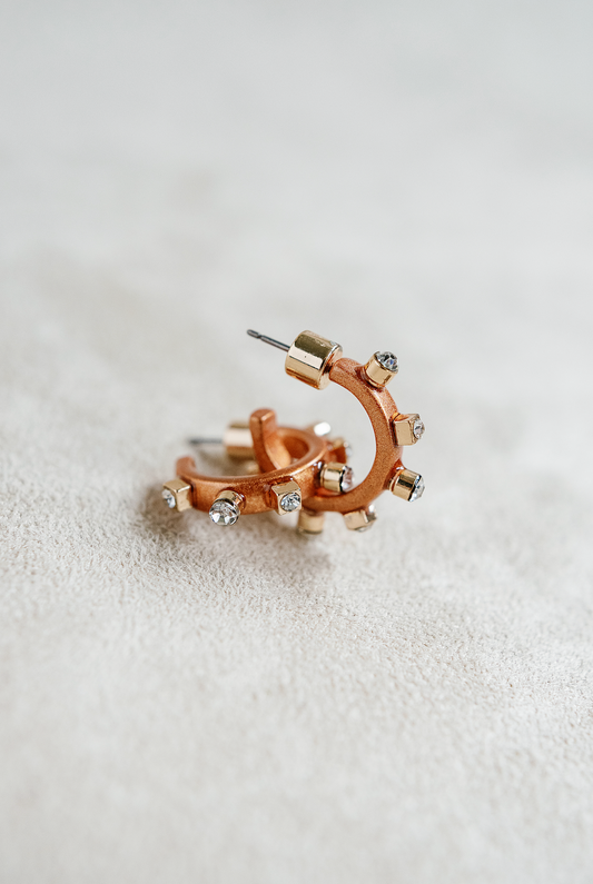A pair of matte metal copper hoop earrings with gold accents, featuring studs around the rim and clear crystals on the post,