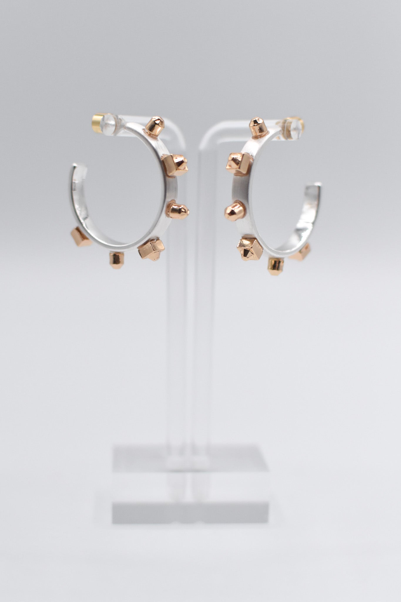 A pair of metallic silver hoop earrings with matte gold studs are mounted on a transparent stand set against a light background.