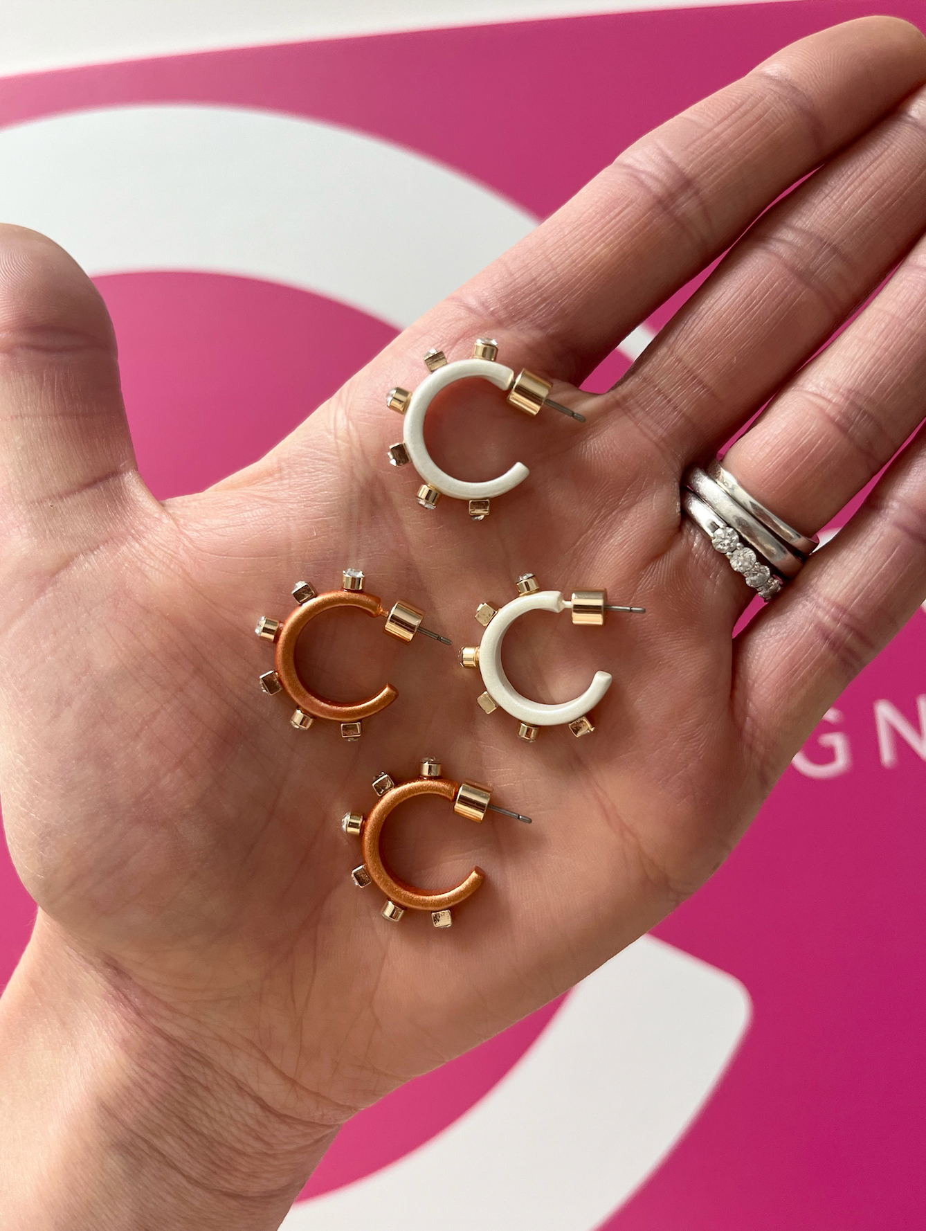 A hand is holding four small hoop earrings in bronze and white, each adorned with studs and lying on a palm in front of a bright pink background.