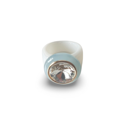 A light blue and white acrylic ring with a round, faceted clear jewel centered in a gold-edged bezel sits against a stark white background.