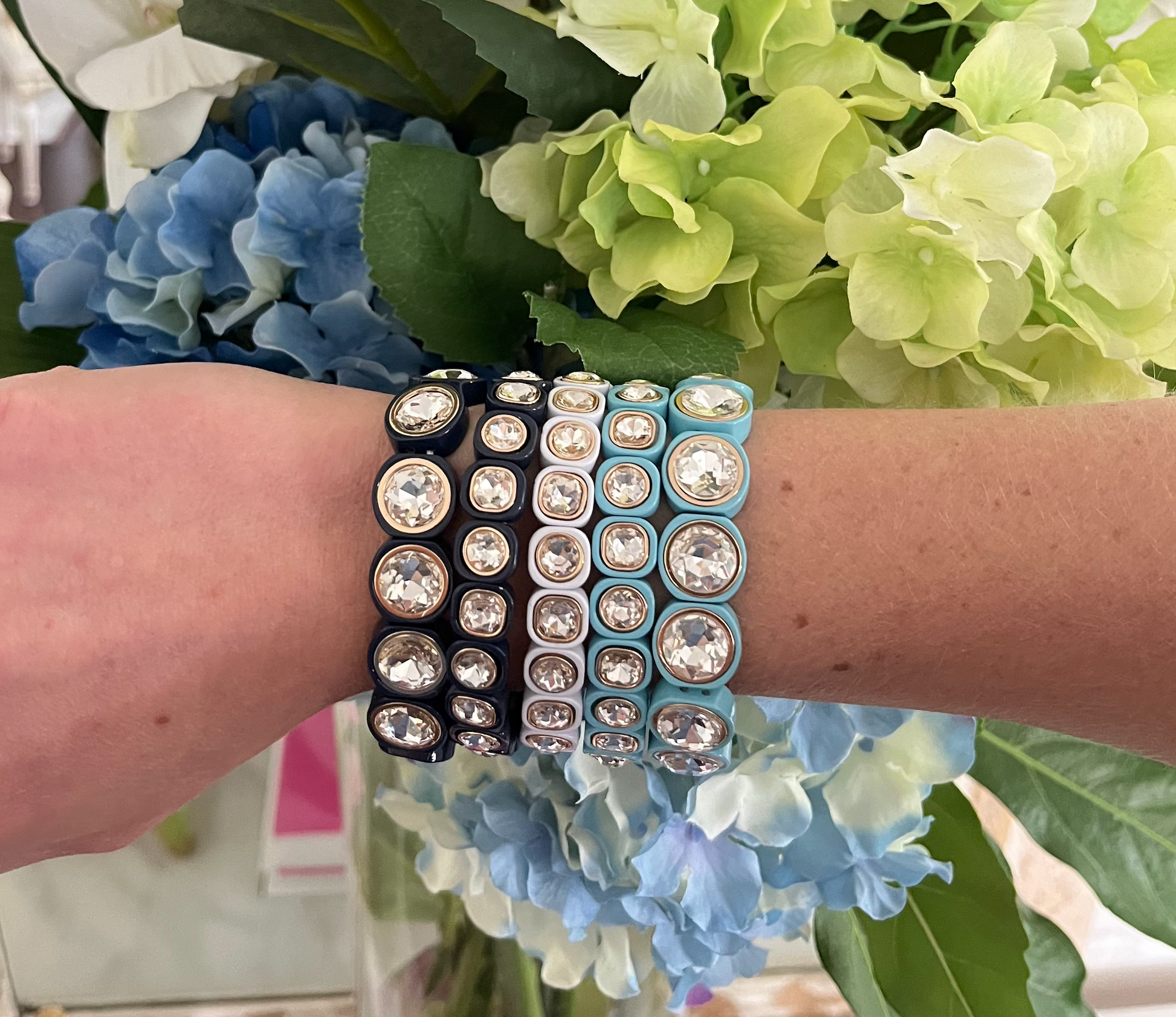 A stack of crystal-studded bracelets in shades of blue, white, and black is displayed on an arm, set against a background of green and blue hydrangeas.