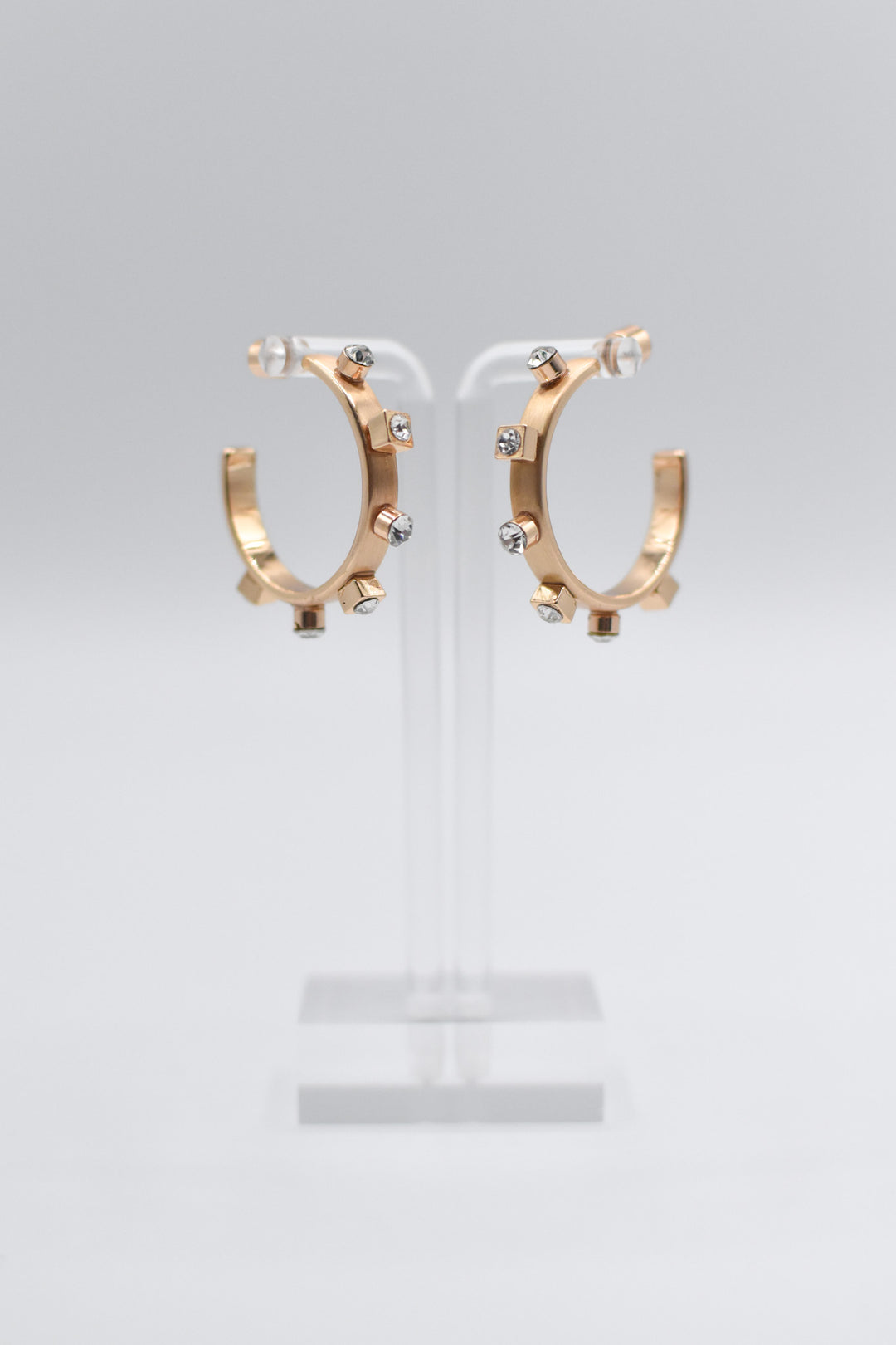 A pair of 14k brushed gold metal hoops, each adorned with six small gold studs and crystal accents, are mounted neatly on a transparent stand.