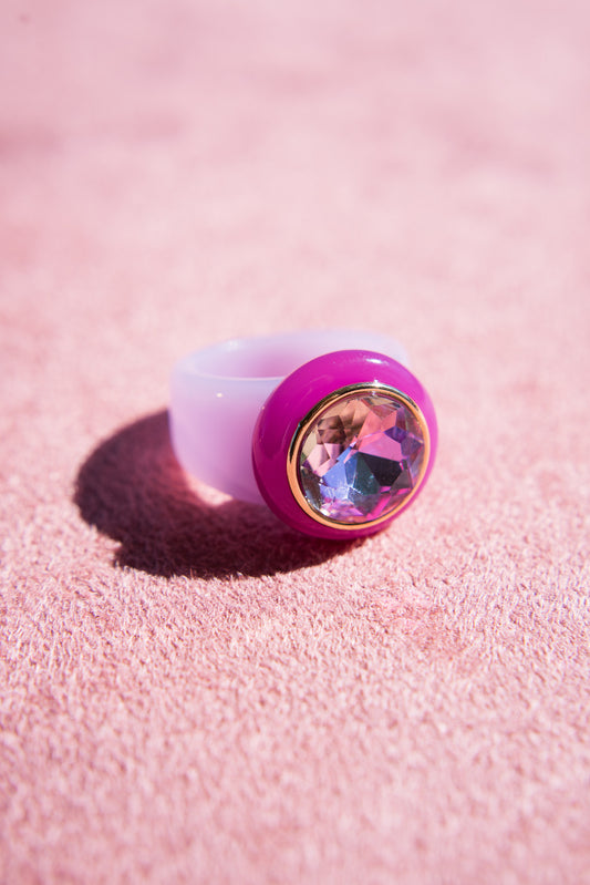 A chunky, light purple acrylic ring with a bold pink bezel, showcasing a large, gemstone reflecting hues of purple and pink, rests on a soft pink surface.