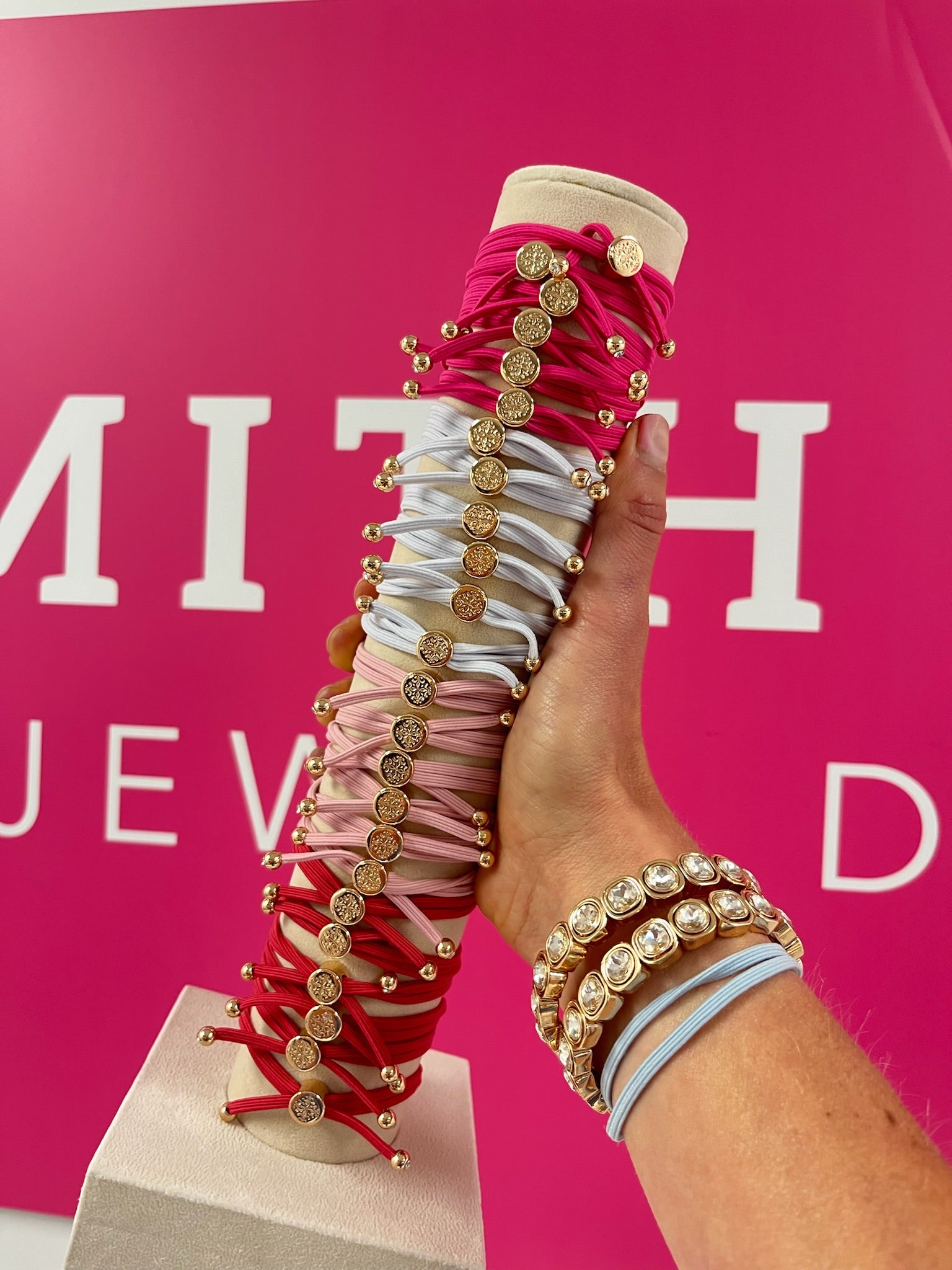 A hand holds stacked hair ties in pinks, reds, and neutrals with gold coin charms, complemented by layered gold-accented crystal bracelets on the wrist.