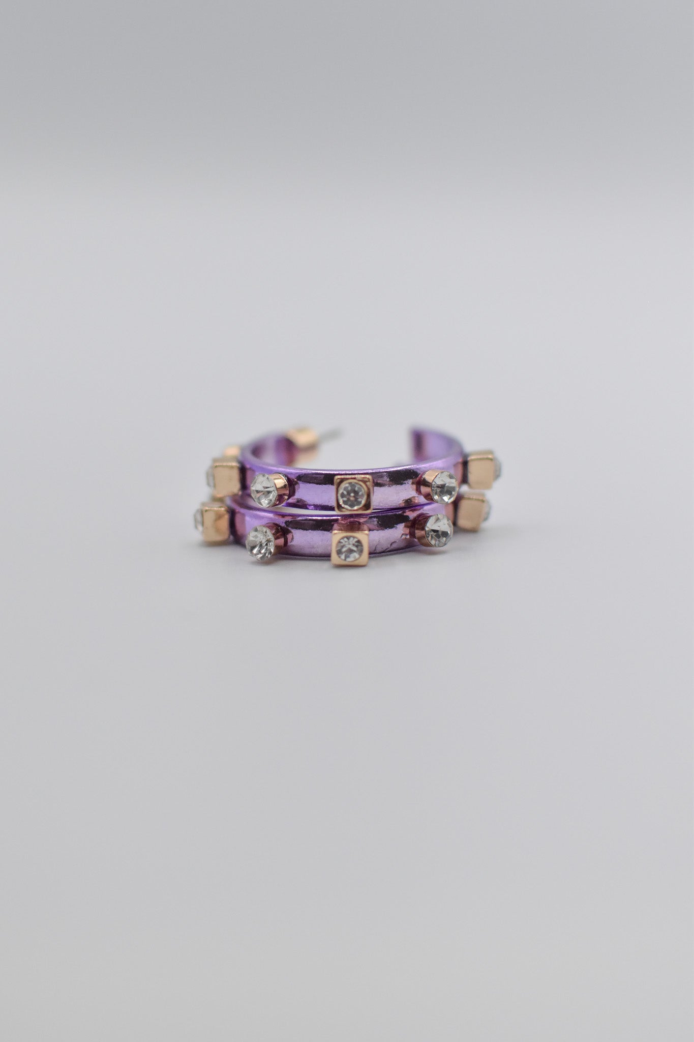 A pair of brushed metallic light purple metal hoops decorated with square and round crystal studs lay stacked neatly on a plain, light-colored background.