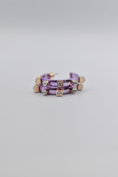 A pair of brushed metallic light purple metal hoops decorated with square and round crystal studs lay stacked neatly on a plain, light-colored background.