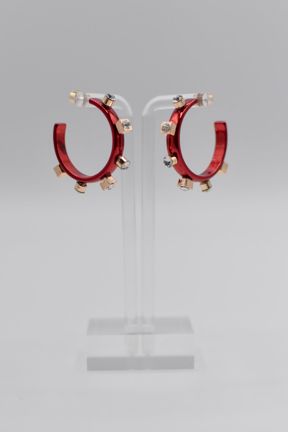 A pair of glossy, petite red metallic metal hoops, each adorned with six small crystal jewels on gold studs, are mounted on a transparent stand.