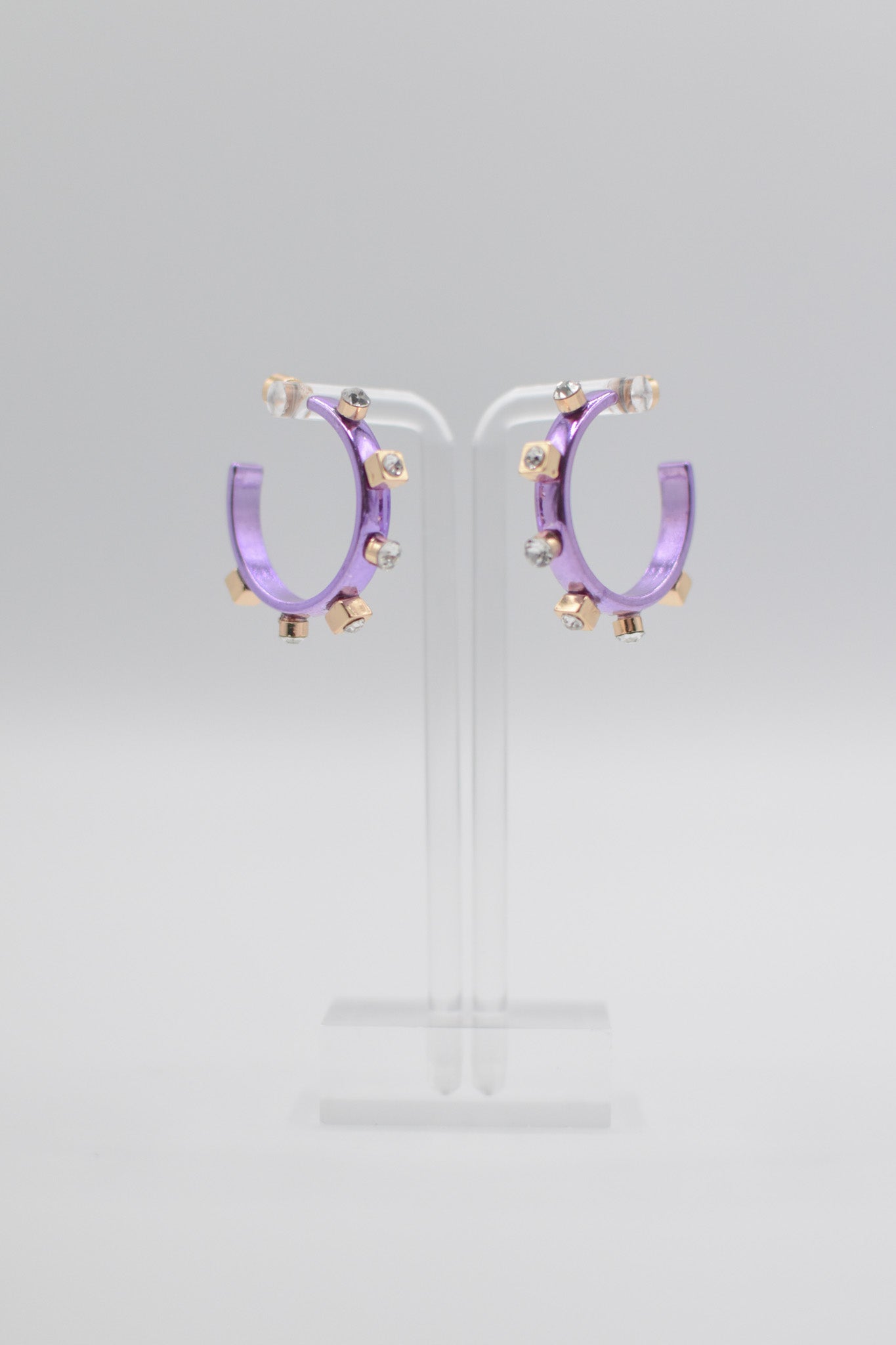 A pair of glossy, light purple metallic metal hoops, each adorned with six small crystal jewels on gold studs, are mounted on a transparent stand.