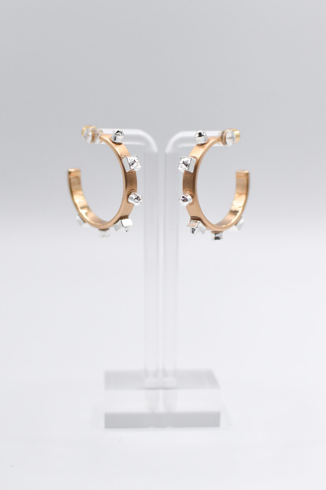 A pair of metallic gold hoop earrings with matte silver studs are mounted on a transparent stand set against a light background.