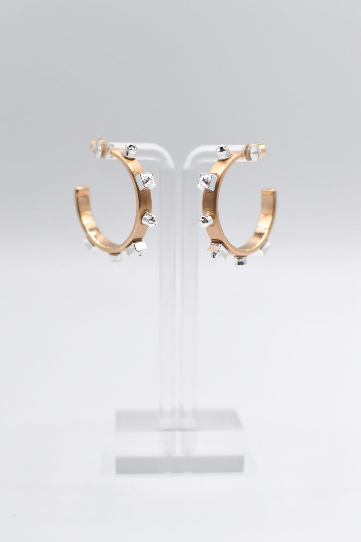A pair of metallic gold hoop earrings with matte silver studs are mounted on a transparent stand set against a light background.