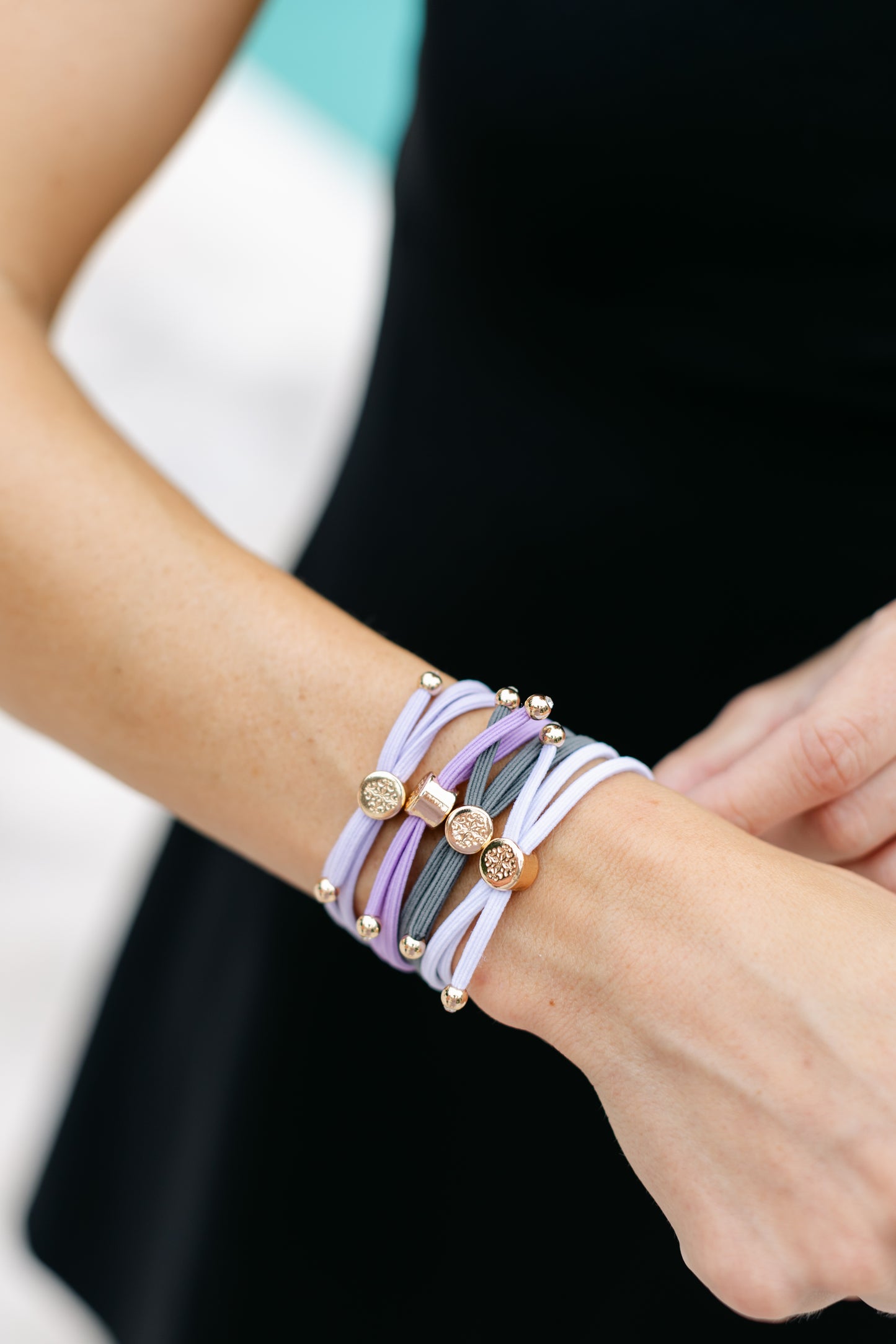 A wrist with multiple stacked elastic bracelets in lavender, grey, and white with gold accents against a black workout top and a blurred background.