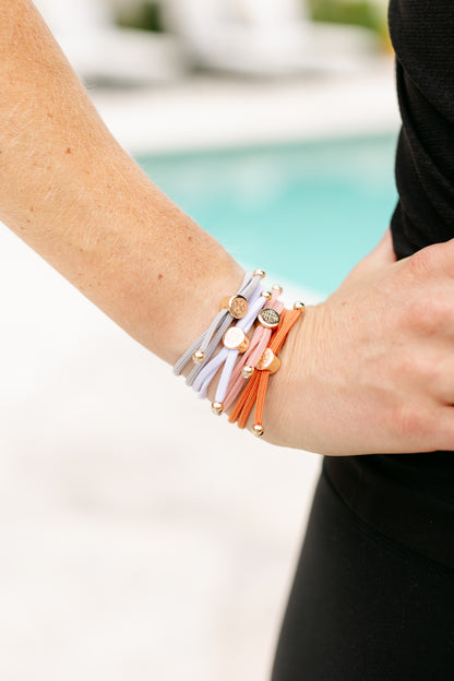 A close-up of a wrist adorned with multiple elastic hair ties in soft gray, white, pink, and orange hues with gold accents, against a poolside.