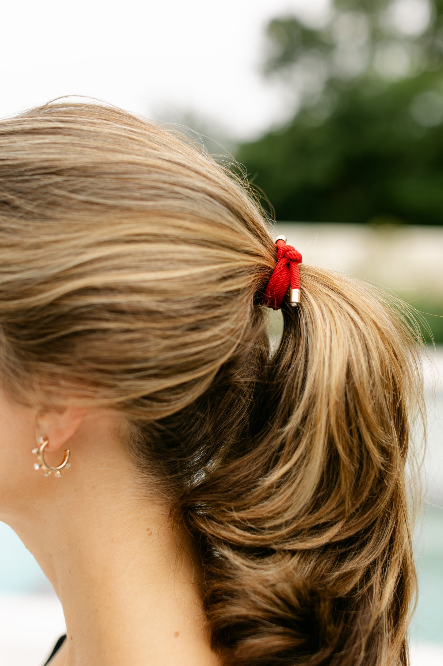 Close-up of a ponytail secured with a bright red elastic hair tie with gold accents, against the model's light brown hair, in an outdoor setting.