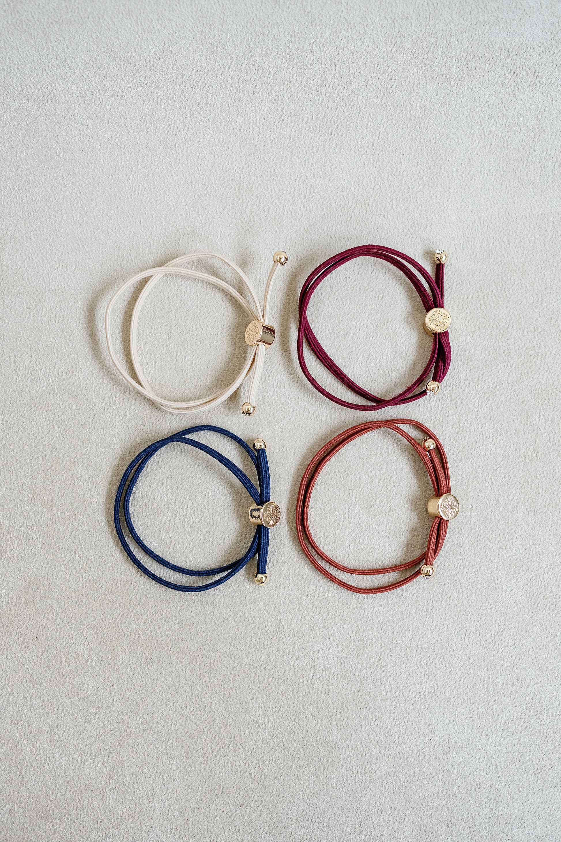 Four neutral-colored elastic hair ties in shades of burgundy, beige, rust, and navy with gold accents arranged in a grid on a white background.