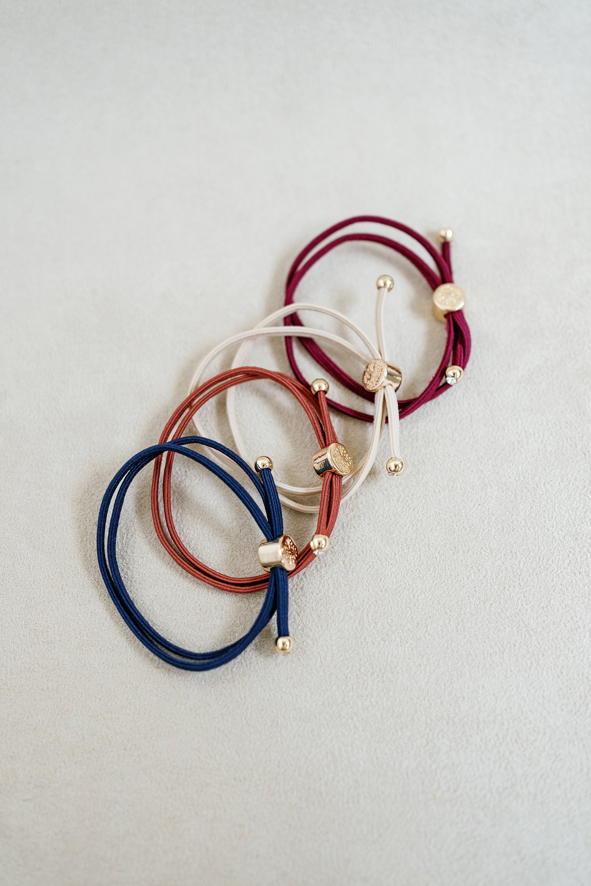 Four neutral-colored elastic hair ties in shades of burgundy, beige, rust, and navy with gold accents arranged in a grid on a white background.