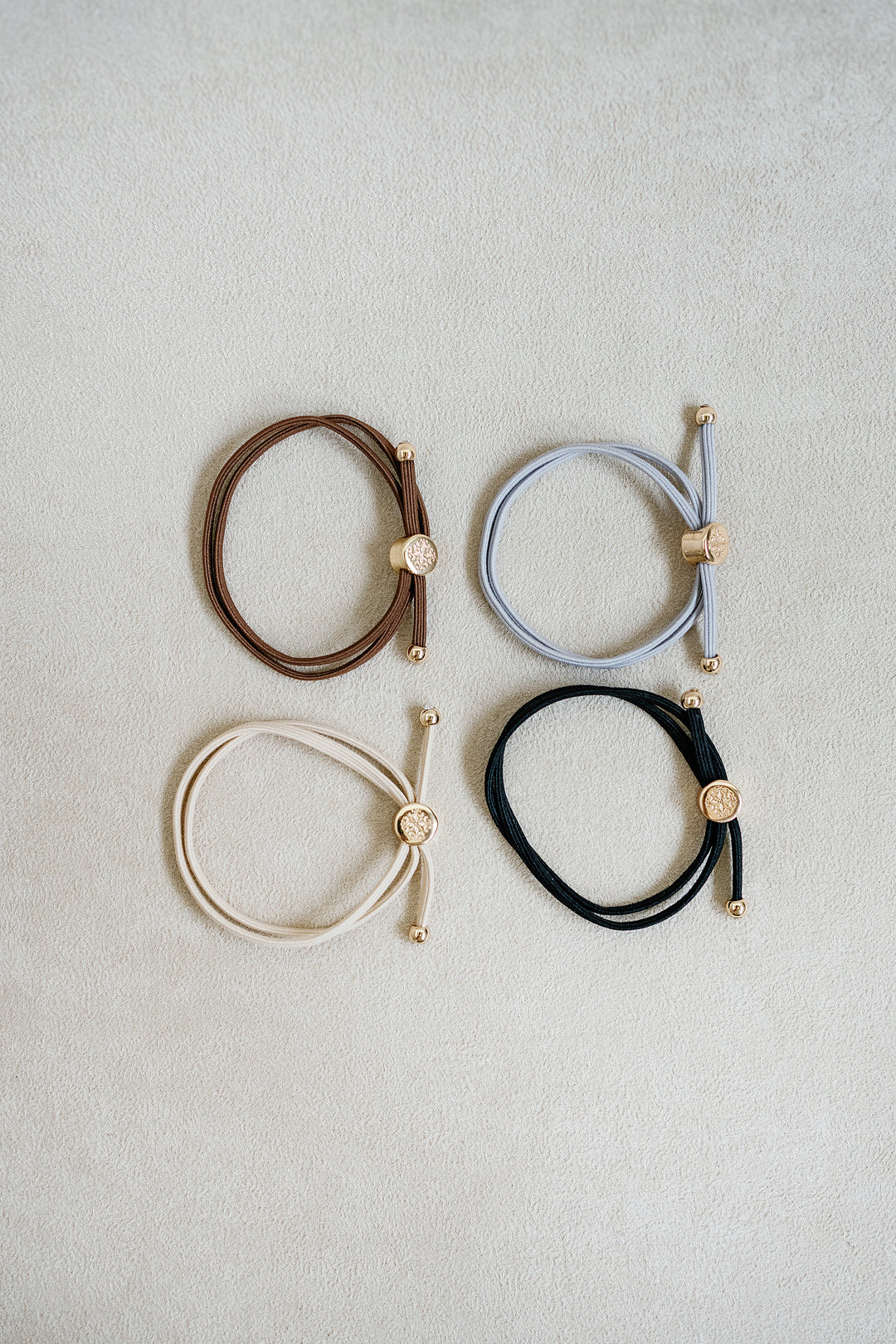 Four neutral-colored elastic hair ties in beige, brown, grey, and black, arranged in a grid with gold accents, showcasing a sleek, minimalist design.