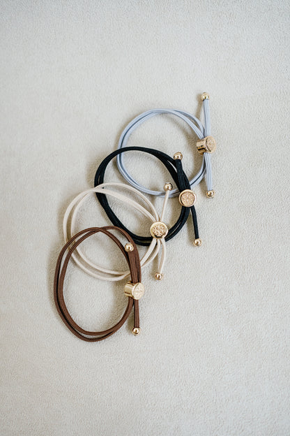 Four neutral-colored elastic hair ties in beige, brown, grey, and black, arranged in a grid with gold accents, showcasing a sleek, minimalist design.