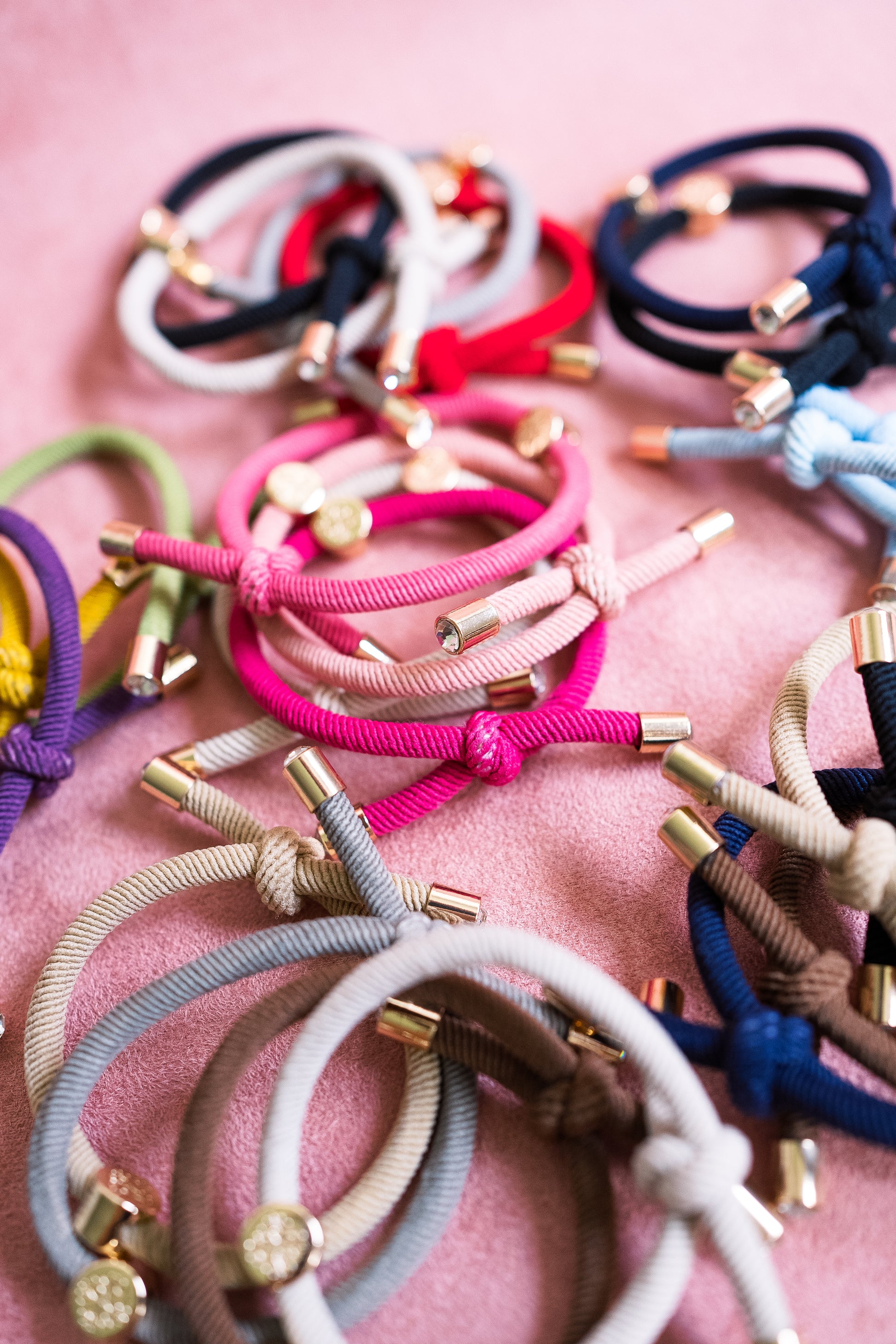 Close-up shot of assorted colorful elastic hair ties with gold accents, arranged on a pink background, featuring shades like pink, navy, green, and beige.