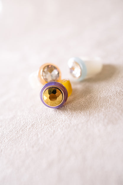 The image features a collection of colorful acrylic rings with large jewel center in various vibrant colors like purple, orange, and blue on a soft fabric.