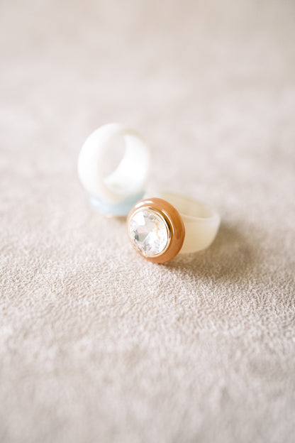 A close-up shot of two acrylic rings, one with a clear gem and gold accent resting on a textured surface, softly lit to highlight the band details.