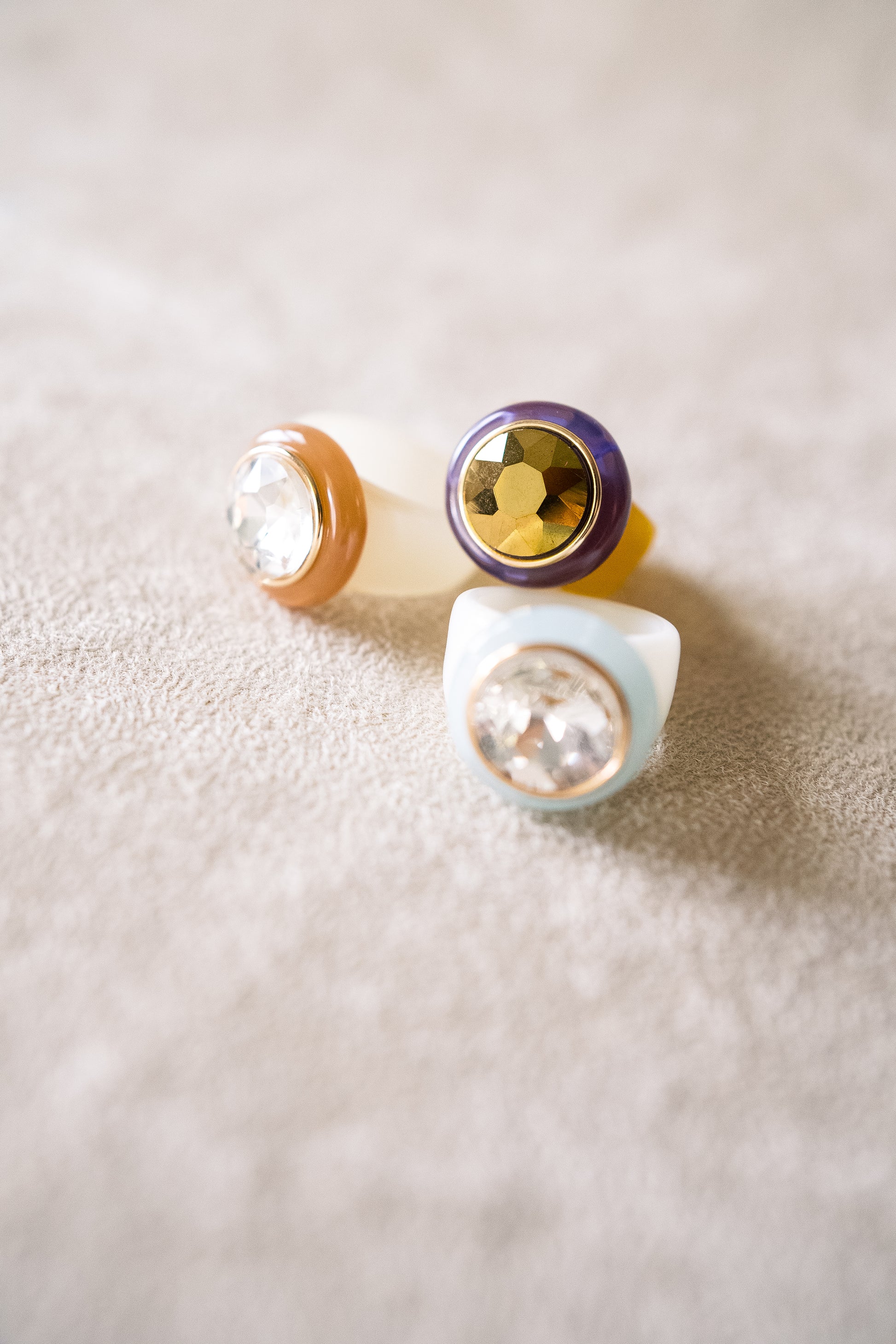 A group of three colorful acrylic rings with faceted, round jewel centers, placed on a soft, textured beige surface.