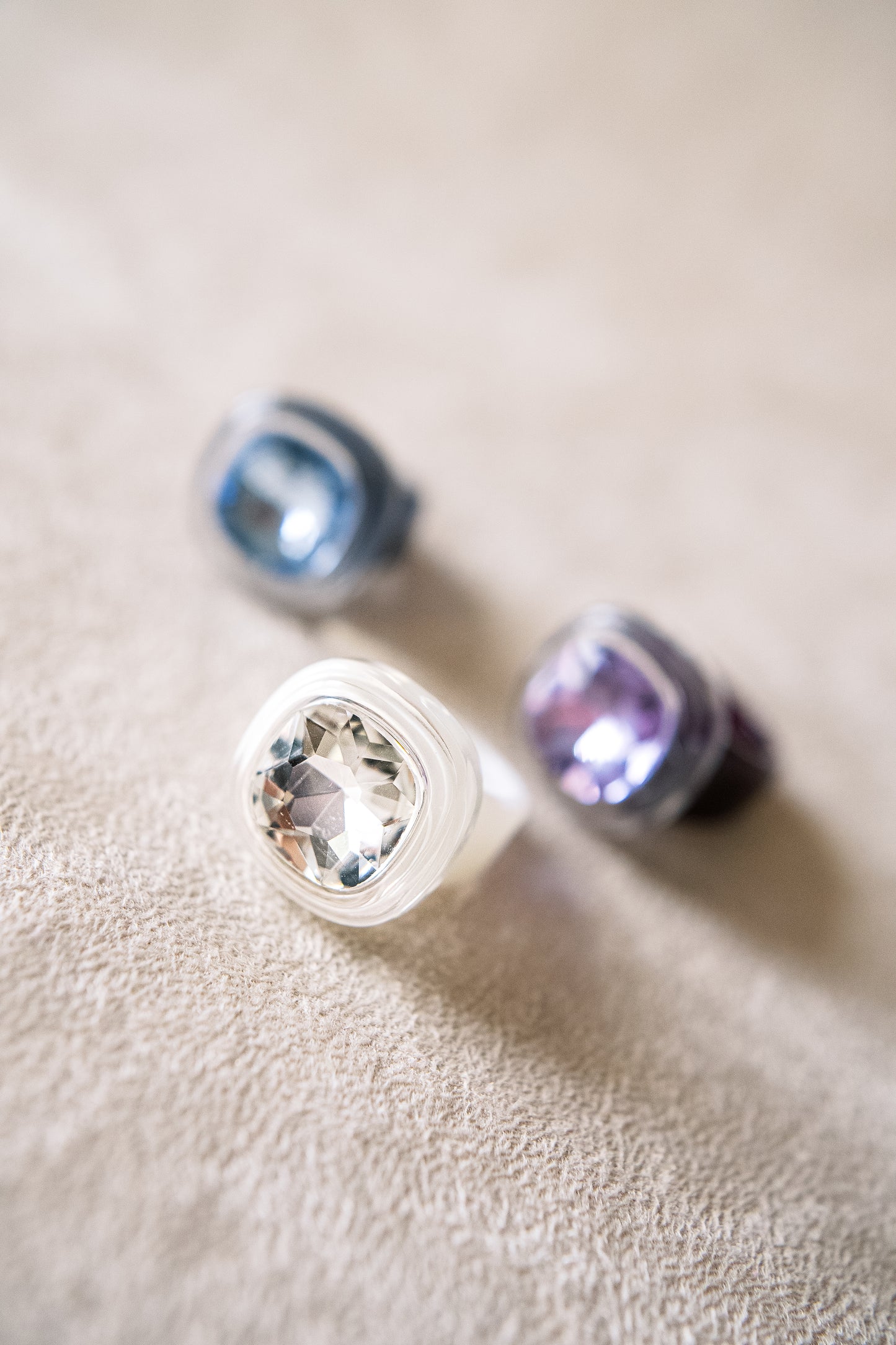 Three acrylic rings in pastel hues of clear, purple, and blue feature faceted gemstones and are arranged on a soft beige surface in focus.