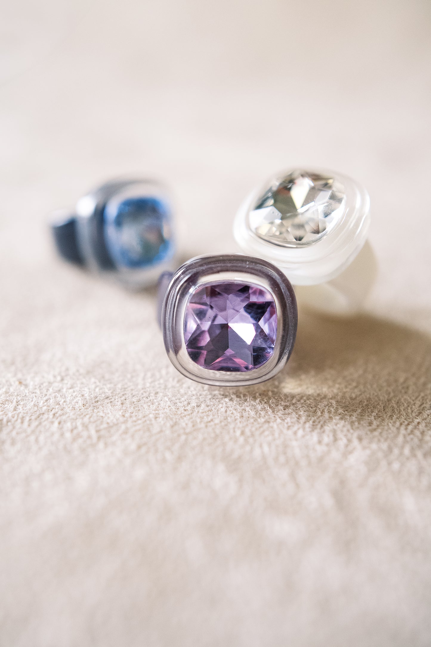 A close-up shot of three rings, each featuring large, faceted gems in clear, purple, and blue tones, set against a soft beige surface for contrast.