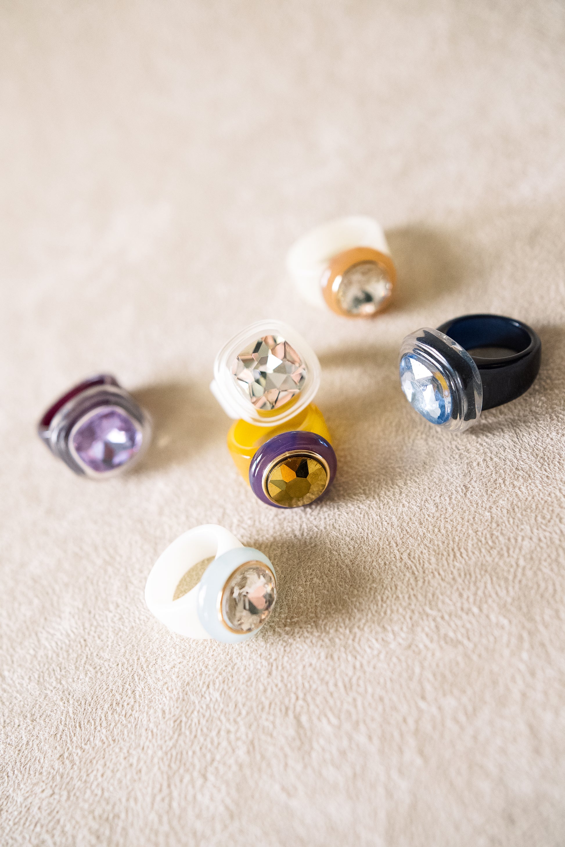 Six acrylic rings with large, faceted jewel centers in various colors—purple, blue, gold, and silver—are scattered on a beige textured surface.