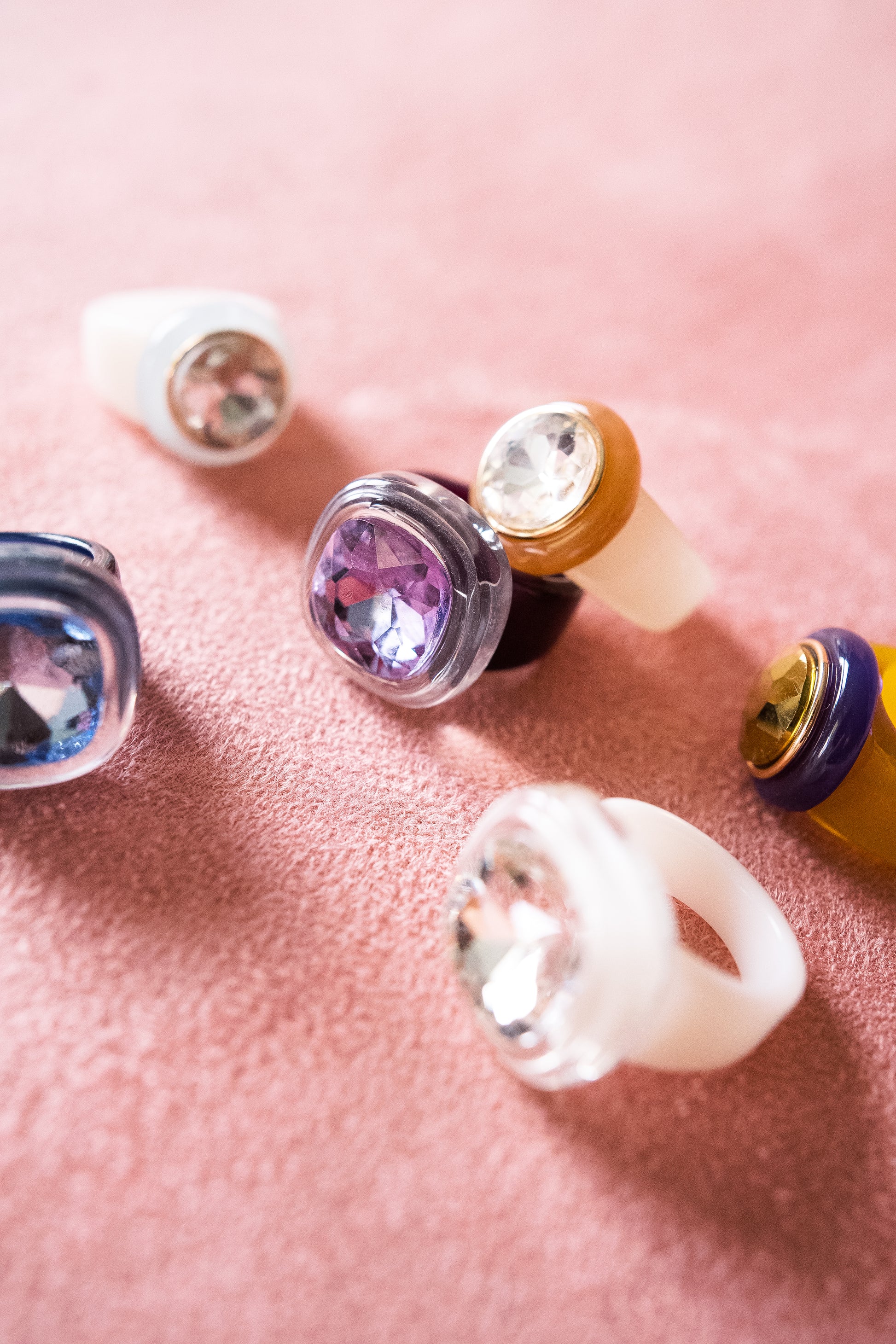 A set of rings rests on a soft, pink textured surface, each featuring a distinct, faceted gemstone in various colors, including purple, blue, and clear.