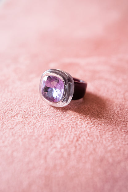 A close-up image of an oval-shaped gemstone ring with a purple-tinted stone set in a purple acrylic bezel, resting on a soft, textured pink surface.