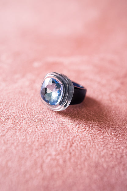 A close-up image of an  oval-shaped gemstone ring with a blue-tinted stone set in a blue acrylic bezel, resting on a soft, textured pink surface.