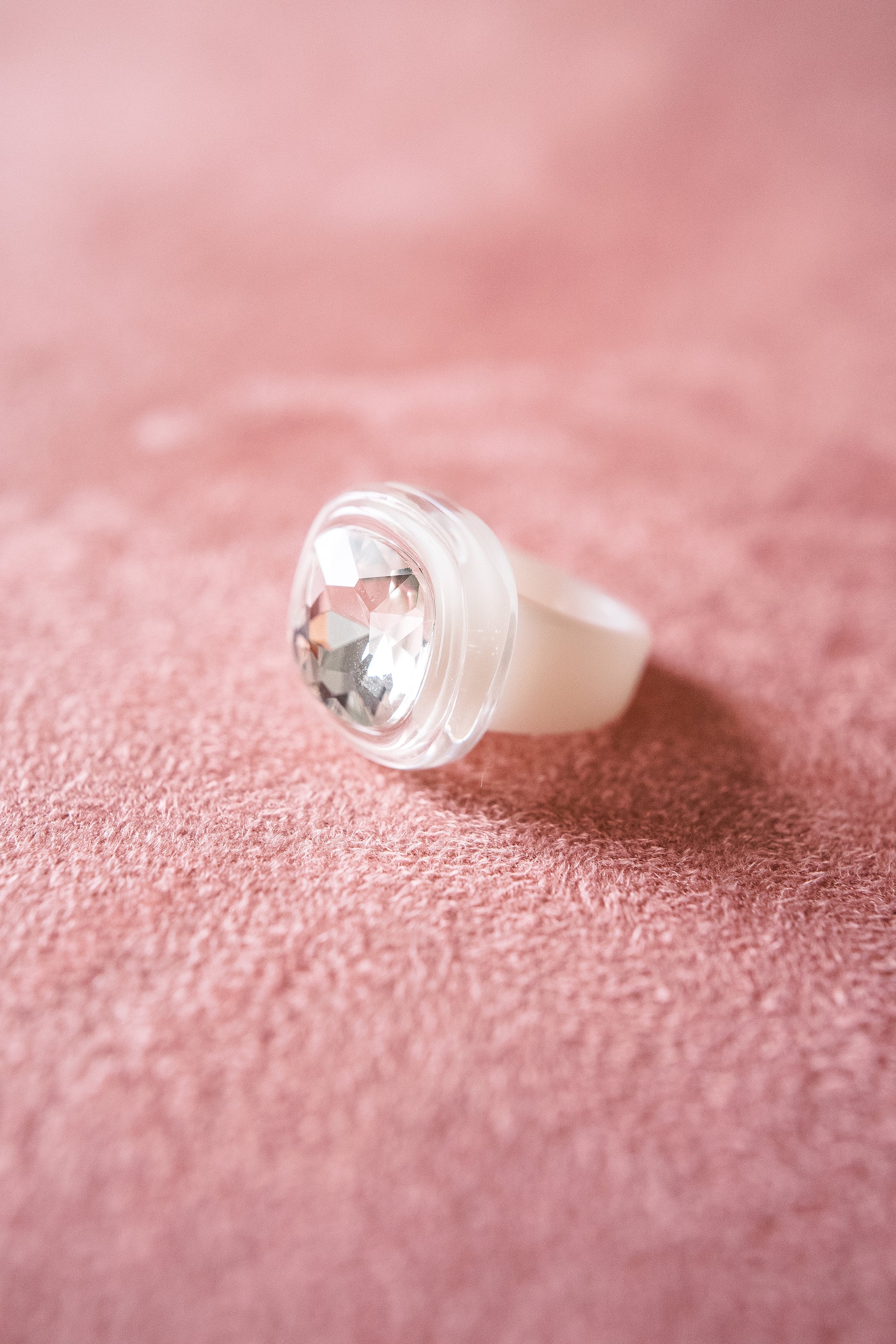 An oversized white acrylic ring with a large clear gemstone is set against  a textured pink surface, highlighting its round smooth surface.