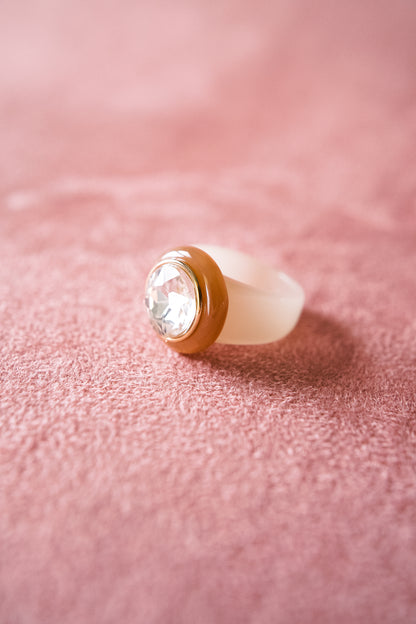 A translucent white ring with a clear round gemstone set in a golden bezel, resting on a soft pink fabric backdrop with a slight warm tone.