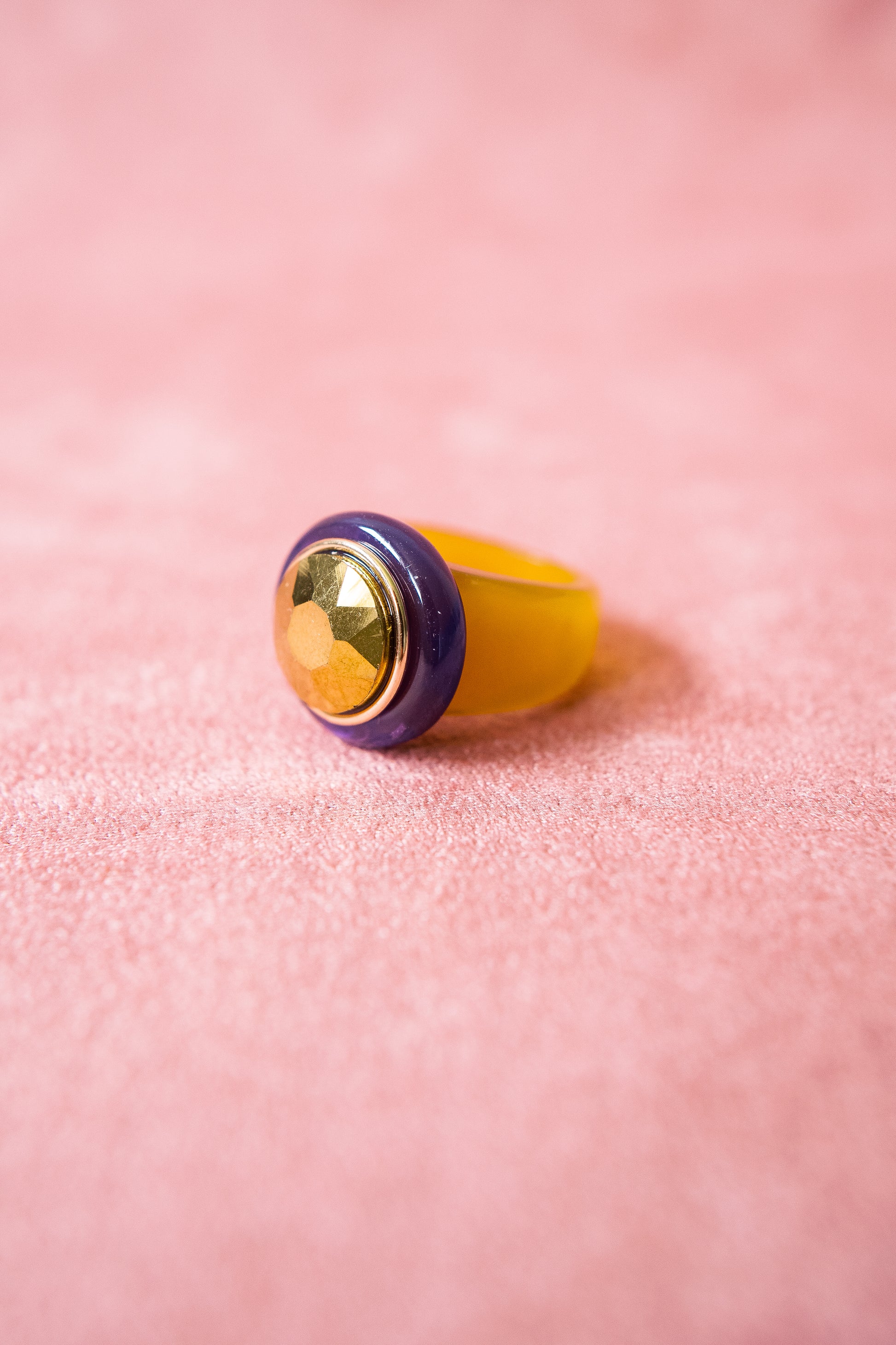 A close-up image of an acrylic ring with a two-tone band in yellow and purple, featuring a gold-accented, faceted center stone reflecting light.