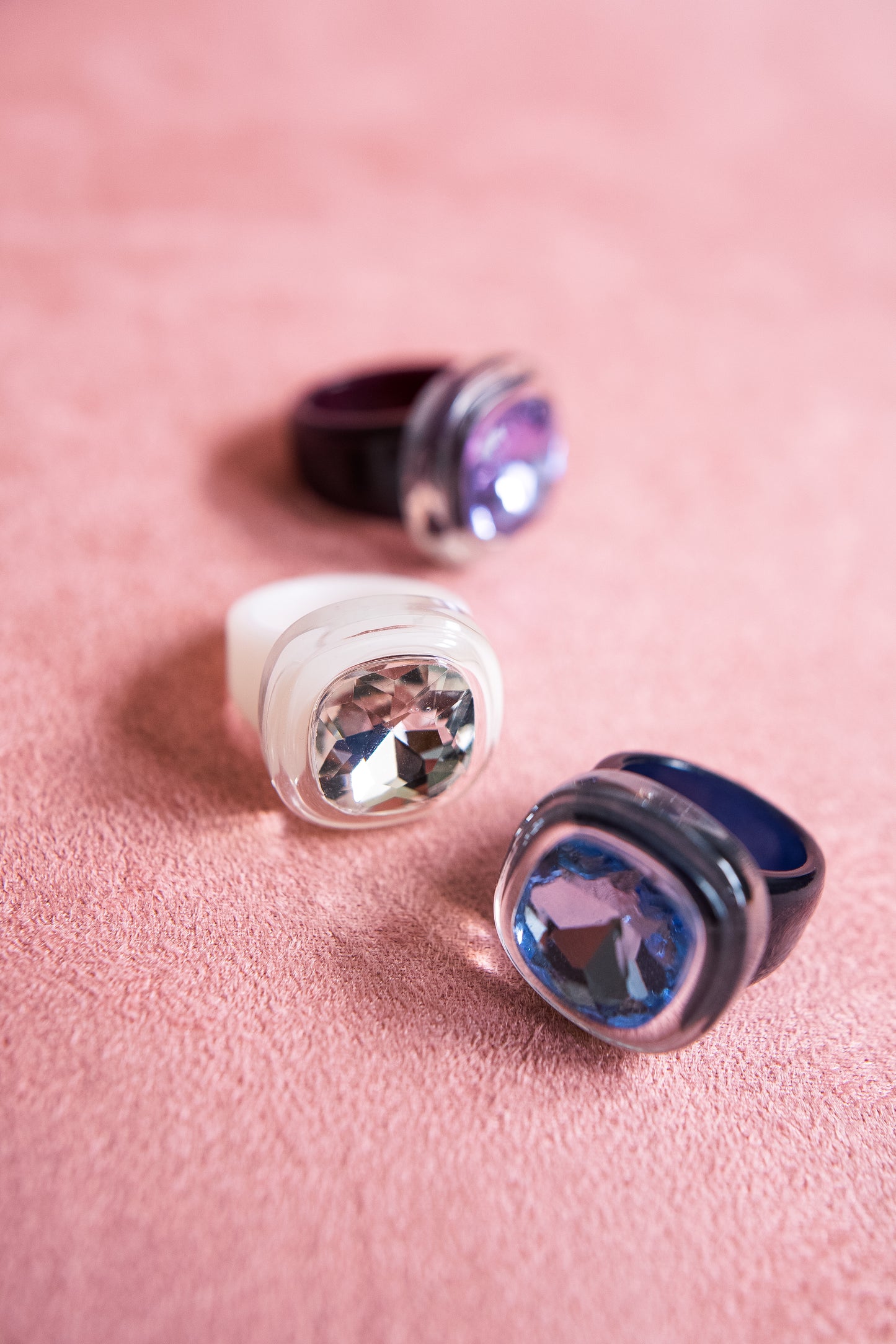 Three chunky rings with large faceted gemstones, placed on a soft pink surface, sparkle in hues of white, purple, and blue from different angles.