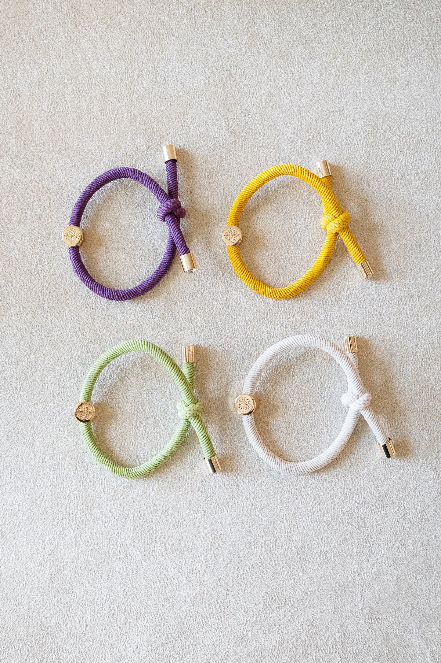 Four elastic hair ties in purple, yellow, green, and white, arranged in a grid on a soft beige surface, each featuring shiny gold accents and knotted designs.